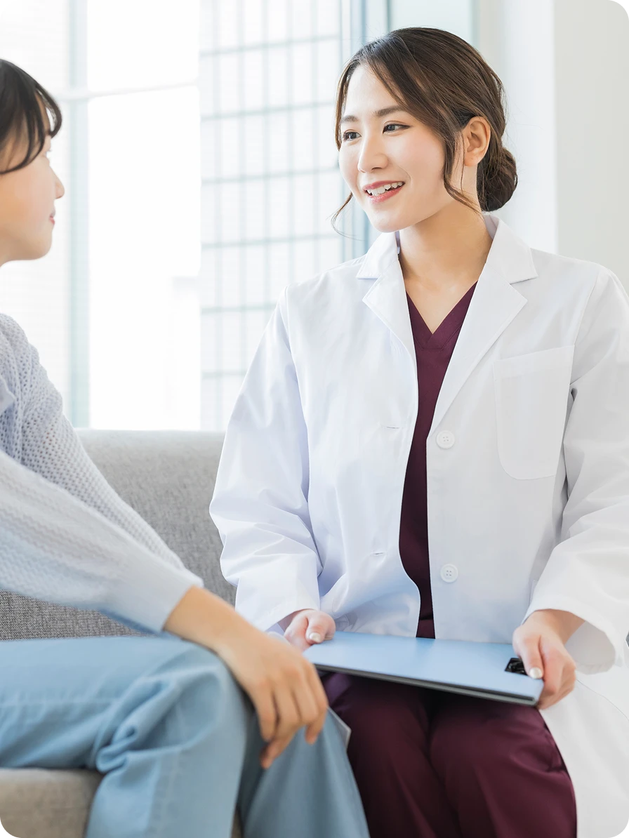 診察室にて、笑顔で温かく⽬の前の患者と向き合う⽩⾐の⼥性医のイメージ